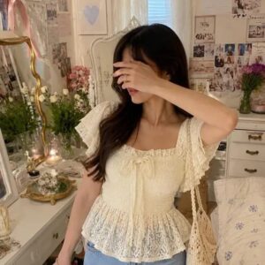 Woman wearing an ivory smocked peplum top with flutter sleeves, a square neckline, and a lace hem